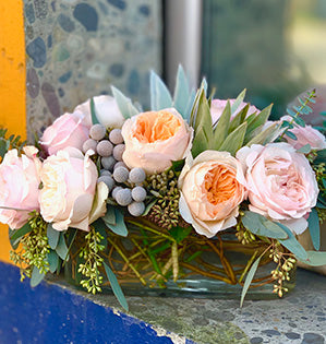 A lovely sweetheart table centerpiece features two kinds of garden roses: Keira (soft pink) and Juliet (peach) along with spiral eucalyptus, seeded eucalyptus, and silver tree protea.