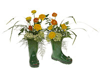 Green rain boots serve as the containers for two floral arrangements of orange and yellow ranunculus, chartreuse green hydrangea, lily grass, plumosa, and Italian ruscus.