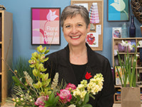 Leanne Kessler stands smiling in the Floral Design Institute's film studio set with an arrangement of Cymbidium orchids, roses, hydrangea, and foliages along with a container of small daffodils. Leanne is wearing a black top and black blazer.