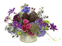 A colorful centerpiece of delphinium, hydrangeas, bachelor buttons, clematis, dusty miller, blueberry roses, astrantia, chocolate lace, purple artichokes, eryngium, and purpke carnations resting in a round galvanized tin container. 