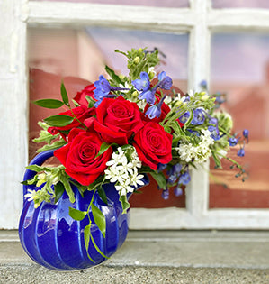 A holiday floral design in a blue Fiestaware pitcher features red roses, delphinium, tweedia, scabiosa, and Italian ruscus.