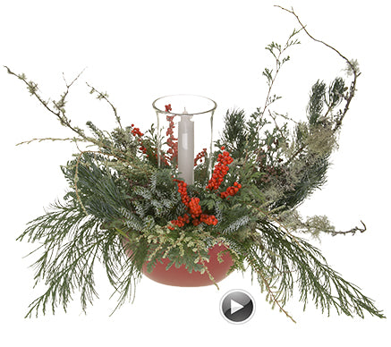 A bespoke style arrangement featuring various conifer foliages, lichen-covered branches, bright ilex berries, with a glass hurricane with white column candle in the center, all in a bright red bowl container.