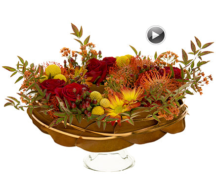 A Thanksgiving centerpiece of vibrant orange protea, bicolor chrysanthemums, vivid red roses, billy balls, yarrow, hypericum, orange-dyed wax flower, and nandina with a base wrapped in magnolia leaves and gold flat wire on a glass platter.