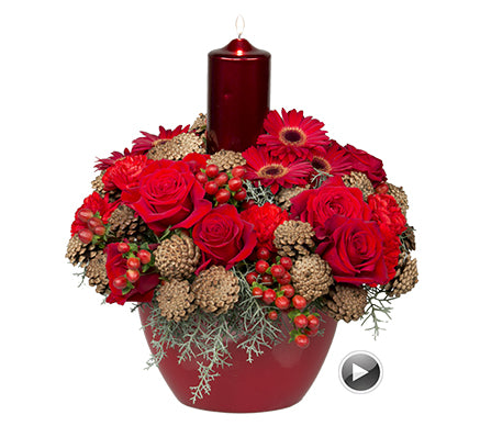 A Christmas centerpiece of red roses, carnations, gerbera daisies, hypericum berries, blue cypress foliage, and pine cones with a shiny red candle emerging from the center in a large red container.