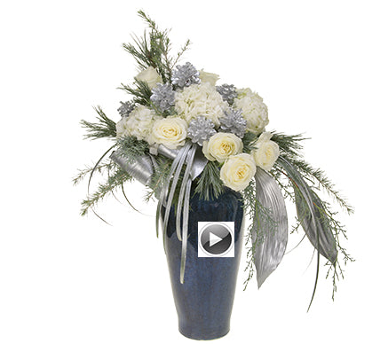 An elevated floral arrangement of blue-based conifer foliage, silver lacquered aspidistra and lily grass, white roses and hydrangea, and silver, sparkly coated pinecones in a tall, blue ceramic vase.