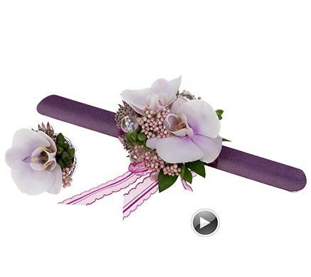 A purple wrist corsage featuring Phalaenopsis orchids, astrantia buds and leaves, rice flower, purple ribbon, and a shiny bead with a coordinating floral ring off to the side.
