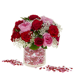 A bold floral arrangement of roses in deep red, playful pink garden roses, and baby's breath in a cylindrical glass vase filled with red, pink, and white M&Ms. Some M&Ms are also strewn about outside of the vase.