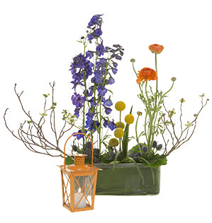 A floral arrangement of delphinium, eryngium, orange ranunculus, billy balls, and blooming branches of young dogwood flowers next to a small orange lantern holding a lit tea candle.
