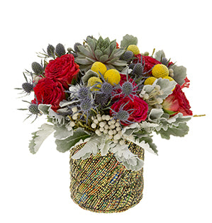 A hand-tied bouquet of red roses, billy balls, and eryngium is gently muted by the grays of accompanying dusty miller, Brunia, and succulent and sits in a large vase.