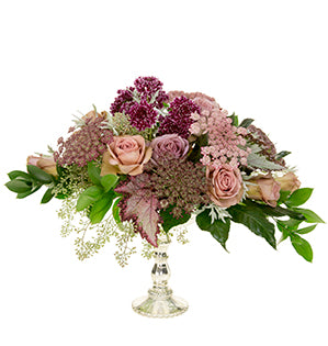 A vintage floral design of Amnesia Rose, silvered foliages, Queen Anne’s Lace in soft pink and burgundy, aubergine Scabiosa and angel wing begonia situated in a mercury glass compote.