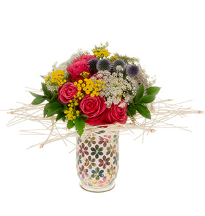 A bouquet of roses, tansy, echinops, Queen Anne's Lace, and ruscus is framed by a collar of midelino triangles rests in a white vase decorated with retro 6-petal flowers in colorful metallics.