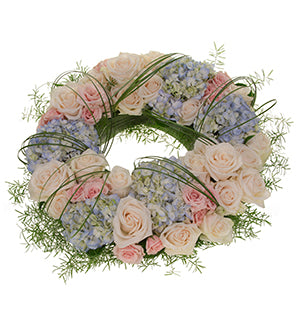 A floral wreath, in soft pinks and blues, filled with hydrangea blossoms, hybrid roses and spray roses. Foliage of Israeli Ruscus and Spring Rye fill out the wreath with sections wrapped in small bundles of Beargrass.