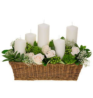 A centerpiece of five white pillar candles sitting at various heights in a bed of green trick dianthus, ruscus, white roses, hypericum berries, and baby's breath sits in a long woven basket.