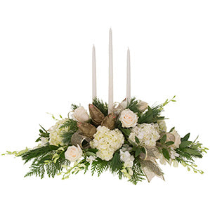 A centerpiece filled with evergreens, bayleaf, white hydrangea, roses, and Dendrobium orchids is accented with gold ribbon and holiday ornaments and holds three tall, skinny white candles.