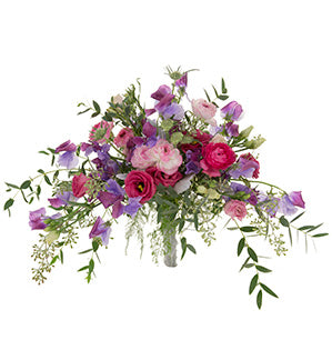 A long, low bouquet combines Sweet Peas, Lisianthus, Ranunculus and Scabiosa buds in shades of pinks and purples with Gunni and seeded eucalyptus in a Dusty Miller wrapped bouquet holder.