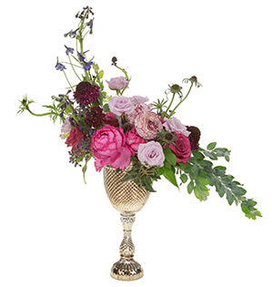 A lavish floral arrangement of roses - Yves Piaget garden, Blueberry standard, and Eloquence spray - Delphinium, viburnum, ranunculus, Scabiosa, and acacia sit atop a silver, tall-footed compote.