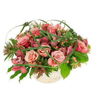 A floral centerpiece in copper tones features copper-hued Toffee Roses, seeded Lily of the Valley bush, Hypericum, Salal, fatsia leaves, and cymbidium orchids. Blades of lily grass are artfully tied and strewn about over the top of the arrangement.