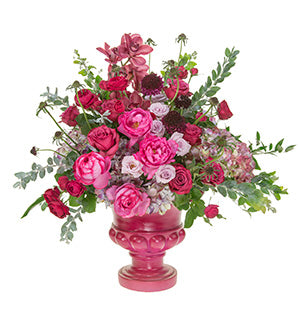 A floral arrangement in pink and purple color tones contains Cymbidium orchids, scabiosa, hydrangea, roses in Garnet Gem, Yves Piaget, Eloquence, and blueberry varieties, Acacia foliage, fatsia leaves, Italian ruscus, and Aspidistra.