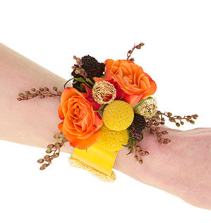 A wrist adorned with a floral wrist corsage of bright orange spray roses, vivid yellow Craspedia, burgundy scabiosa buds, and little pieris seed bods in burgundy, with gold wire balls and a yellow band.