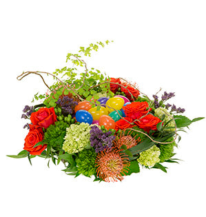 A springtime nested floral centerpiece of red roses, hydrangea, green trick dianthus, orange pincushion protea, hypericum berries, Italian ruscus, lily grass, galax leaves, curly willow, and maidenhair fern holds colorful plastic Easter eggs.