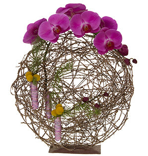 A loosely-woven standing disc is topped with Phalaenopsis orchids, enhanced with Australian foliages, and holds bundles of Billy balls wrapped in pink bullion wire.