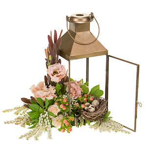 A brass candle lantern appears to be overflowing with lily of the valley, hypericum berries, leucadendron, peach lisianthus, galax leaves, and mini carnations. A bird's nest and eggs are also inside the lantern.