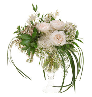 A floral bouquet featuring crisp white garden roses accompanied by hydrangeas, miniature cymbidiums, pieris, wax flowers, and foliages including Sprengeri, salal, Italian ruscus, Israeli ruscus, and lily grass draping down from the sides.