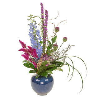 A tall arrangement in linear design contains liatris, delphinium, misty, bunny tail, celosia, hydrangea, fatsia leaves, lily grass, and curly willow sits in a round, blue vase.