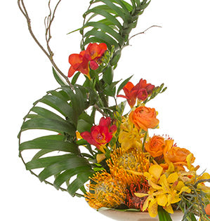 A unique woven palm nautilus is a perfect addition to a beautiful floral design that combines roses, orchids, protea, and reflexed tulips in sunset hues.