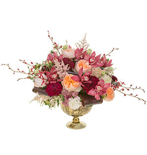A floral centerpiece in a glamorous metallic gold compote bursts with Juliet garden roses, lisianthus, cymbidium orchids, dahlias, astilbe, James Storie orchids, and astrantia, all in shades of blush and burgundy, with folded ti leaves at the base.