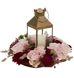 A brushed gold candle lantern holds a white tower candle and sits inside a round tray filled with pink roses, burgundy dahlias, heather, and verbena.