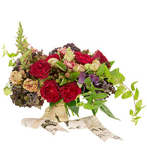A hand-tied bespoke garden style bouquet containing blush lisianthus, antique carnations, snap dragons, Tess garden roses, berries, grains, hydrangea, and fatsia are tied with a thick patterned ribbon.