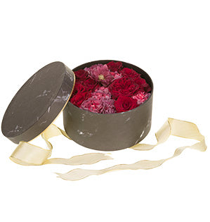 An open hat box has a black marble-looking covering and is filled with Black Baccara Roses, Antique Bi-Colored Carnations and Hellebores. The lid is leaning on the box and is lined at the tip with gold foil, and a gold mesh ribbon lays strewn about.