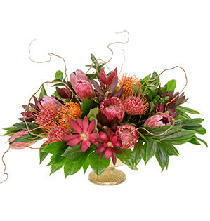 A compote style floral centerpiece in dark pink, deep mahogany and orange hues is created with proteas, leucadendron, cocculus, Israeli ruscus, fatsia, and pittosporum.