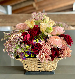 An autumn basket floral design mixes chrysanthemums, spray roses, ranunculus, and lisianthus in deep red and dusty pink colors.