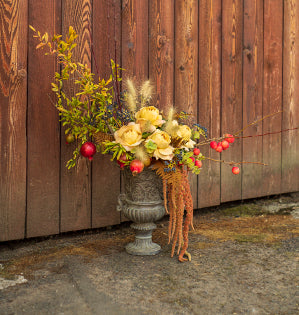A lush floral design rests in a detailed urn and mixes crabapples and pomegranates on their branches to enhance the garden roses, privet berries, and dried ferns.