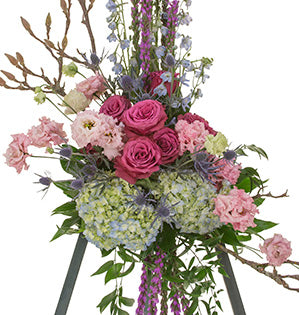 A large easel spray consists of light blue hydrangeas, dark pink roses, blossoming spring branches, blush carnations, purple liatris, blue bells, and Italian ruscus.