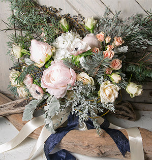 A gorgeous hand tied bouquet features pale pink peonies, cream roses, peach spray roses, white hydrangea, dusty miller, evergreens, and other foliages, all tied off with an ivory satin ribbon.