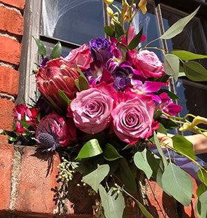 A beautiful wedding bouquet in jewel tones features pink roses, purple tulips, coral protea, orchid blossoms, blue eryngium, seeded eucalyptus, and salal.