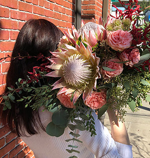 This lush and elegant wedding bouquet mixes blush king protea, coral garden roses, marsala orchids, and three kinds of eucalyptus: seeded, gunni, and silver dollar.