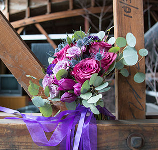This gorgeous boho style wedding bouquet, creThis gorgeous wedding bouquet mixes cherry garden roses, pink spray roses, eryngium, red and purple tulips, silver dollar eucalyptus, and is finished with purple sheer and lavender satin ribbons.