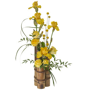 A dramatic vertical arrangement in bright yellow featuring bearded iris, roses and craspedia with lily grass, galax, and Italian ruscus foliages sitting in a bamboo container.