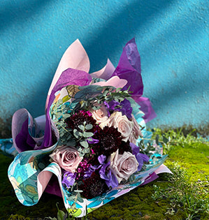 A beautiful hand tied bouquet is filled with violet blooms including roses, scabiosa, and eucalyptus, then it is wrapped in colorful waterproof papers to enhance its look.