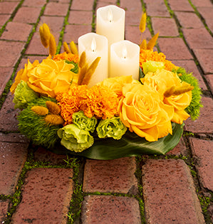A beautiful circular floral design with candles in the center mixes Creme Brulee roses, marigolds, green trick dianthus, lisianthus, and bunny tails.