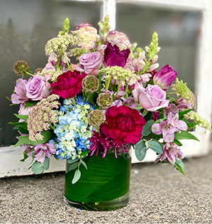 A classic vase of flowers in a pink, red and lavender color palette is created by mixing roses, carnations, ranunculus, and tulips with scabiosa pods and Queen Anne's lace. 
