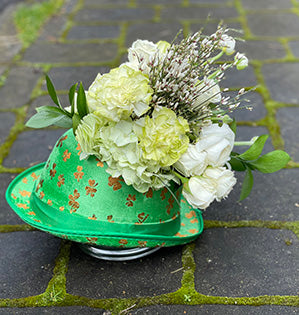 A festive holiday arrangement features white hydrangeas, spray roses, carnations in a green St. Patrick's party hat