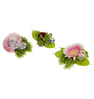 A trio of boutonnieres: one with a soft pink camellia against green trick dianthus with a spiral of silver wire, another with a small succulent and a jewel, the third with more green trick dianthus and a micro gerbera.