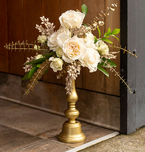 A beautiful elevated centerpiece features Princess Miyuki roses, Blanche spray roses, aspidistra, fatsia, Israeli ruscus, gold eucalyptus, and faux gold foliage.