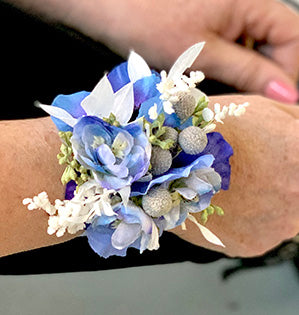 A beautiful wrist corsage combines dried and bleached materials with silk flowers in purple, blue and white colors.