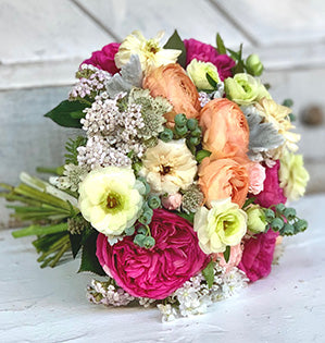 A gorgeous wedding bouquet in a peach and pink color palette mixes lovely luxury materials like garden roses, ranunculus, spray roses, blueberries, hypericum, lamb’s ear, and rosemary.